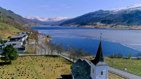 Aerial-shot-of-a-snowy-lake-next-to-a-green-valley-with-a-church-with-a-weather-vane-and-a-cemetery
