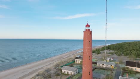 Aerial-establishing-view-of-red-colored-Akmenrags-lighthouse,-Baltic-sea-coastline,-Latvia,-white-sand-beach,-calm-sea,-sunny-day-with-clouds,-wide-drone-shot-moving-forward
