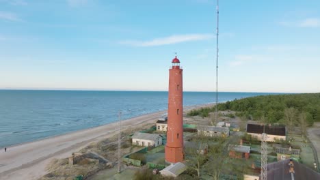 Aerial-establishing-view-of-red-colored-Akmenrags-lighthouse,-Baltic-sea-coastline,-Latvia,-white-sand-beach,-calm-sea,-sunny-day-with-clouds,-wide-drone-shot-moving-forward,-tilt-down