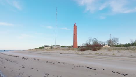 Aerial-establishing-view-of-red-colored-Akmenrags-lighthouse,-Baltic-sea-coastline,-Latvia,-white-sand-beach,-calm-sea,-sunny-day-with-clouds,-low-wide-drone-shot-moving-forward