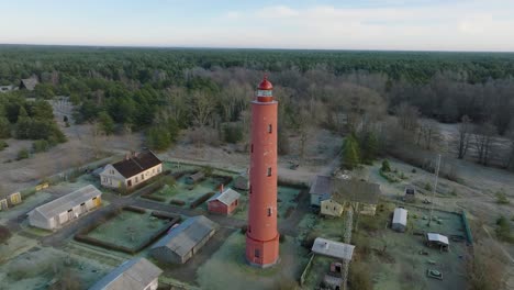 Aerial-establishing-view-of-red-colored-Akmenrags-lighthouse,-Baltic-sea-coastline,-Latvia,-white-sand-beach,-calm-sea,-sunny-day-with-clouds,-distant-wide-drone-orbit-shot