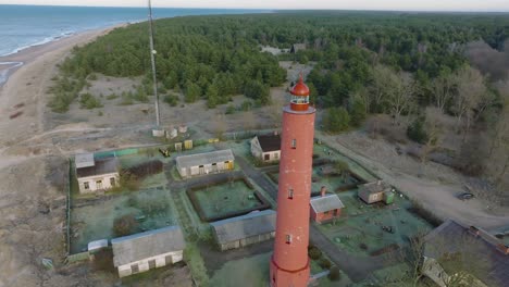 Aerial-establishing-view-of-red-colored-Akmenrags-lighthouse,-Baltic-sea-coastline,-Latvia,-white-sand-beach,-calm-sea,-sunny-day-with-clouds,-wide-drone-orbit-shot