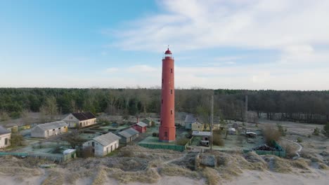 Aerial-establishing-view-of-red-colored-Akmenrags-lighthouse,-Baltic-sea-coastline,-Latvia,-white-sand-beach,-calm-sea,-sunny-day-with-clouds,-wide-ascending-drone-shot-moving-forward