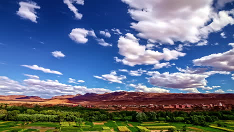 Timelapse-of-a-landscape-of-red-earth-and-mud-buildings-next-to-green-vegetation-under-a-blue-sky-with-advancing-white-clouds