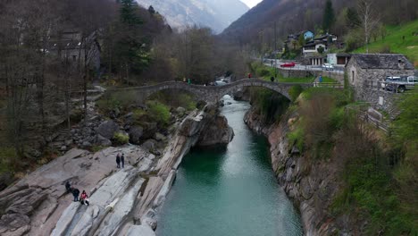Luftaufnahme-über-Einem-Türkisfarbenen-Fluss-Und-Einer-Alten-Und-Malerischen-Brücke-Im-Val-Verzasca-In-Der-Schweiz