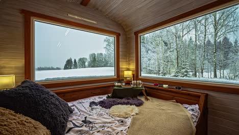 Inside-View-Of-Bed-In-Corner-Of-Rustic-Wood-Cabin-With-Large-Windows-With-View-Of-Snow-Winter-Landscape-Forest-Outside