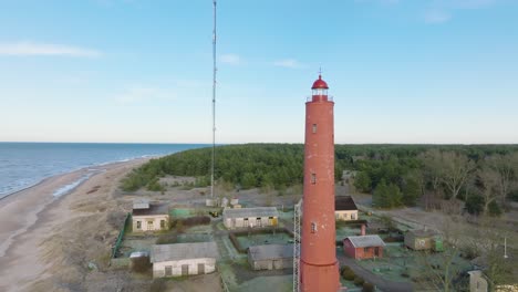 Aerial-establishing-view-of-red-colored-Akmenrags-lighthouse,-Baltic-sea-coastline,-Latvia,-white-sand-beach,-calm-sea,-sunny-day-with-clouds,-wide-ascending-drone-shot-moving-forward