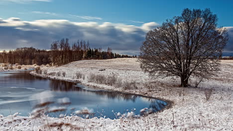 Timelapse-View-Of-Sunny-Snow-Winters-Day-Of-Clouds-Rolling-Past-Being-Reflected-In-Riverbank