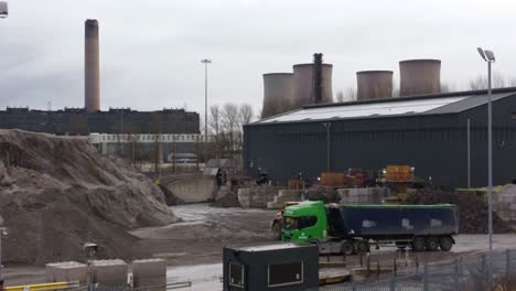 Bulldozer-loading-coal-ash-into-dumper-truck-under-power-station-industrial-skyline-aerial-orbiting-view