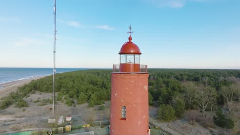 Aerial-establishing-view-of-red-colored-Akmenrags-lighthouse,-Baltic-sea-coastline,-Latvia,-white-sand-beach,-calm-sea,-sunny-day-with-clouds,-medium-ascending-drone-orbit-shot