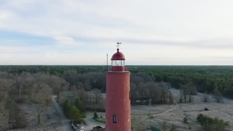 Aerial-establishing-view-of-red-colored-Akmenrags-lighthouse,-Baltic-sea-coastline,-Latvia,-white-sand-beach,-calm-sea,-sunny-day-with-clouds,-wide-drone-shot-moving-backward