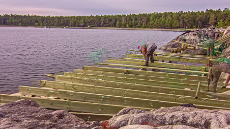 Men-workers-building-wooden-frame-on-rocky-lake-coastline,-fusion-time-lapse