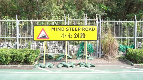 Yellow-Warning-Road-Sign-Stating-Mind-Steep-Road-With-Cyclist-Rising-Past-In-Tseung-Kwan-O,-Hong-Kong