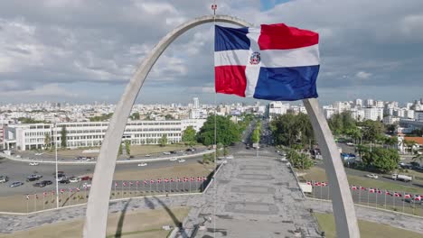 Bandera-De-La-República-Dominicana-Ondeando-En-La-Plaza-De-La-Bandera-Con-La-Ciudad-De-Santo-Domingo-En-El-Fondo-En-República-Dominicana