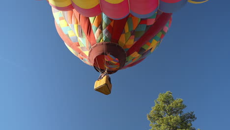 Colorful-Hot-Air-Balloon-With-People-in-Basket-Going-Up-Under-Blue-Sky,-Low-Angle-View,-Parachute-and-Burner-Flames