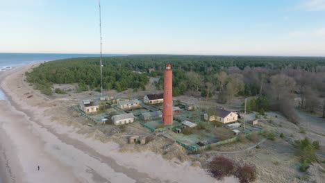 Aerial-establishing-view-of-red-colored-Akmenrags-lighthouse,-Baltic-sea-coastline,-Latvia,-white-sand-beach,-calm-sea,-sunny-day-with-clouds,-wide-distant-drone-orbit-shot
