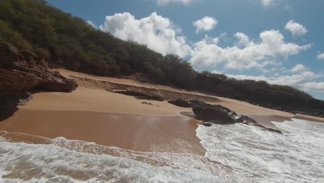 FPV-drone-shot-low-over-a-sunlit-beach-and-over-waves-on-the-coast-of-Hawaii,-USA