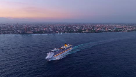 Costa-Pacifica-Cruise-ship-exiting-Santo-Domingo-port-at-sunset-with-coast-and-cityscape-in-background,-Dominican-Republic