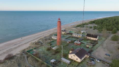 Aerial-establishing-view-of-red-colored-Akmenrags-lighthouse,-Baltic-sea-coastline,-Latvia,-white-sand-beach,-calm-sea,-sunny-day-with-clouds,-wide-drone-shot-moving-forward