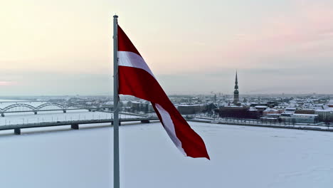 Orbiting-aerial-view-of-a-Latvian-flag-waving-in-the-wind-over-a-snowy,-winter-city-and-frozen-river