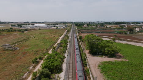 Static-aerial-footage-of-a-red-passenger-train-passing-underneath-in-Bari,-Italy