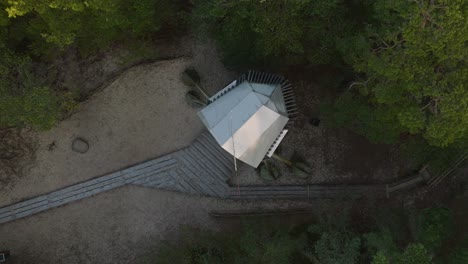 Aerial-birdseye-view-of-modern-boat-shaped-observation-watchtower-in-the-middle-of-pine-tree-forest,-Nordic-woodland,-forest-trail,-sunny-evening,-golden-hour-light,-ascending-drone-shot