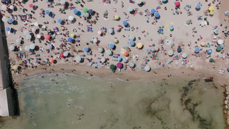 Top-down-aerial-footage-moving-away-from-tourists-on-the-beach-with-an-assortment-of-colors-in-Bari,-Italy-with-the-waves-of-the-sea-moving-in