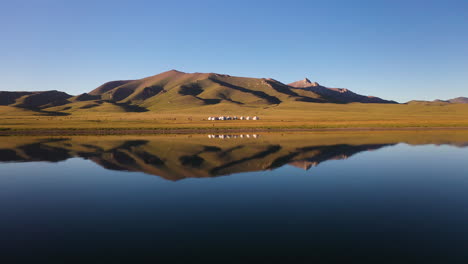 Epic-drone-shot-of-a-small-yurt-village-beside-the-Song-Kol-lake-in-Kyrgyzstan,-with-reflection-on-the-water
