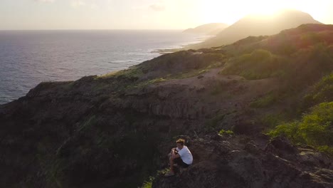 Man-resting-on-top-of-a-cliff-at-sunset-hiking-in-Makapuu-Hawaii---AERIAL-DOLLY-BACK-TILT