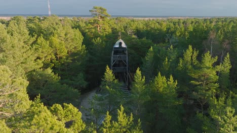 Aerial-establishing-view-of-modern-boat-shaped-observation-watchtower-in-the-middle-of-pine-tree-forest,-Nordic-woodland,-forest-trail,-sunny-evening,-golden-hour-light,-drone-shot-moving-forward