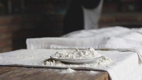 Spilled-Flour-And-Baking-Cloth-On-A-Wooden-Table-With-Woman-Baker-In-The-Background
