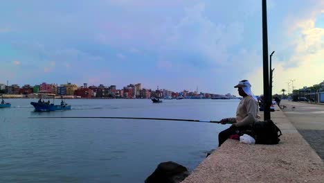 Boats-Passing-by-on-a-Cloudy-Day-as-an-Egyptian-Fisherman-is-Angling-with-a-Long-Fishing-Rod-in-Ras-El-Bar-where-the-Nile-River-meets-with-the-Mediterranean-Sea