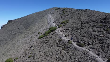 Hiker-walking-up-a-trail-towards-the-peak-of-a-rocky,-barren-mountain