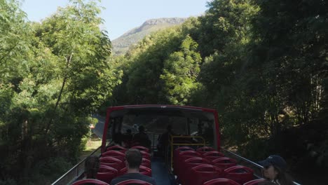 Open-double-decker-tour-bus-drives-narrow-curving-road-in-trees,-POV