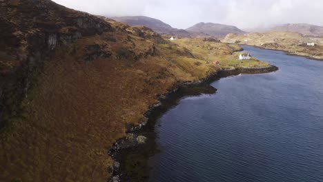 Drone-shot-of-a-collection-of-remote,-coastal-houses-with-a-Scottish-mountain-range,-moorland-and-peatland-in-the-background
