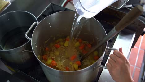 Chef-pouring-a-cup-of-water-into-a-big-stock-pot-full-of-potatoes,-carrots-and-onions-on-stovetop,-chef-cooking-in-the-kitchen,-over-the-shoulder-view