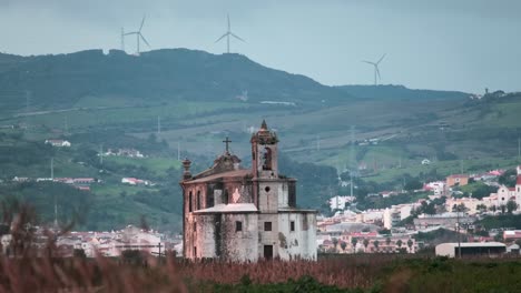 Church-"Ermida-de-Nossa-Senhora-de-Alcame"-on-the-background-of-the-city-of-Lisbon,-green-mountains-and-rotating-wind-turbines-with-wheat-in-the-foreground-at-sunset