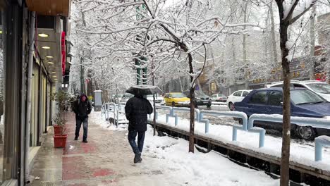 Ein-Alleiniger-Mann-Mit-Schwarzem-Kleid-Hält-Einen-Regenschirm-In-Einer-Komplett-Weißen-Landschaft-Im-Schnee,-Schneeflocken,-Gehweg,-Gefrorene-Straßenbahnen,-Die-Menschen-Fahren,-Die-In-Teheran,-Iran,-In-Der-Wintersaison-Spazieren-Gehen