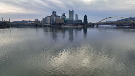 Aerial-glide-over-Ohio-River-with-Pittsburgh-skyline,-Point-State-Park,-and-yellow-bridges-in-background