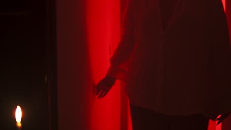 Slow-motion-cinematic-shot-of-a-person-in-a-white-blouse-stroking-a-wall-with-the-hand,-walks-forward-in-an-environment-with-red-lighting-and-burning-candles