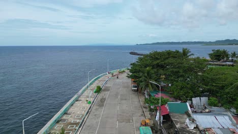 Aerial-Overhead-Drone-shot-of-Road-on-Seawall-between-beautiful-ocean-and-small-barangay-in-Catanduanes,-Philippines