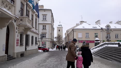 Men,-women-and-children-in-winter-clothing-walk-along-snow-covered-streets-surrounded-by-idilic-buildings-an-a-clock-tower