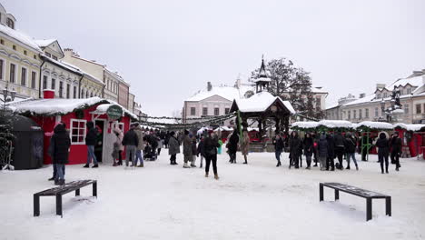 Men,-women-and-children-attend-a-Christmas-fair-of-festive-stalls-in-a-snow-covered-town-square-surrounded-by-idilic-buildings