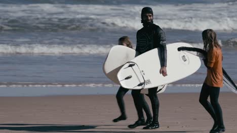 Familia-De-Surfistas-Llevando-Surf-Y-Caminando-Por-La-Playa,-Océano-De-Fondo,-Día-Soleado