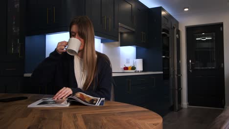 female-model-in-a-blue-cardigan,-bolero-top-drinking-from-a-cup-reading-a-magazine-and-looking-at-phone-at-a-kitchen-table-in-a-modern-kitchen
