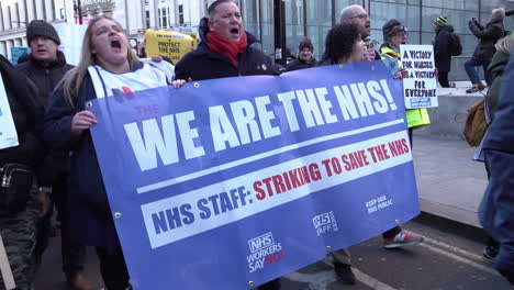 Mitarbeiter-Des-Nationalen-Gesundheitsdienstes-Und-Marschieren-Mit-Einem-Banner-Mit-Der-Aufschrift-„Wir-Sind-Die-NHS.“