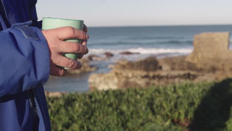 Person-holding-cup-of-tea-or-coffee-outdoors,-ocean-coast-in-background,-copyspace-for-titles