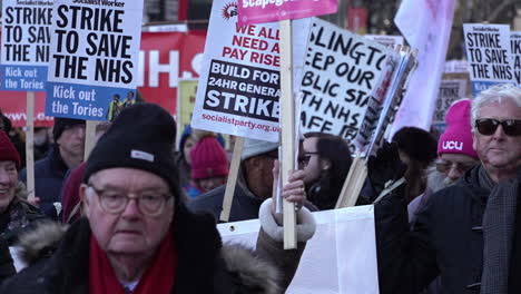 In-Zeitlupe-Schließen-Sich-Unterstützer-Streikenden-Mitarbeitern-Des-Nationalen-Gesundheitsdienstes-An-Und-Marschieren-Mit-Plakaten,-Um-Für-Höhere-Löhne-Und-Bessere-Arbeitsbedingungen-Zu-Protestieren