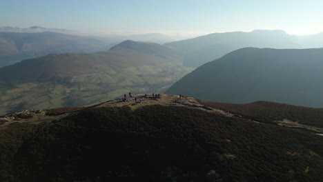 Hikers-on-mountain-summit-with-pull-back-reveal-of-green-valley-and-panoramic-view-of-misty-mountains-beyond-in-English-Lake-District-UK
