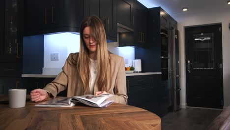 female-model-in-a-smart-jacket-drinking-from-a-cup-reading-a-magazine-and-looking-at-phone-at-a-kitchen-table-in-a-modern-kitchen